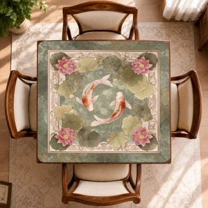 May include: A square table cover featuring a central design of two koi fish swimming in a pond surrounded by green lily pads and pink lotus flowers. The border is decorated with an intricate cream-colored pattern on a muted green background.