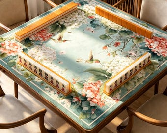 May include: A square mahjong table featuring a light blue surface decorated with white and pink hydrangea flowers and hummingbirds. Four rows of orange and white mahjong tiles are arranged along the edges of the table, surrounded by four wooden chairs with light-colored upholstered seats.