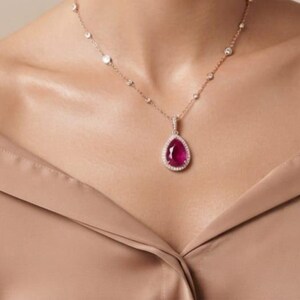Könnte beinhalten: Eine Halskette aus Roségold mit einem tropfenförmigen, dunkelrosa Edelsteinanhänger, der von einem Kranz aus kleinen weißen Steinen umgeben ist. Die zarte Kette ist mit gleichmäßig verteilten runden weißen Steinen besetzt, was ein raffiniertes Schmuckstück ergibt.