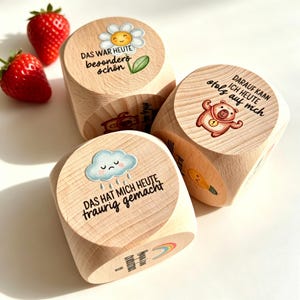 Könnte beinhalten: Drei helle Holzwürfel mit aufgedruckten Illustrationen und deutschem Text. Ein Würfel zeigt ein Gänseblümchen mit 'DAS WAR HEUTE besonders schön', ein weiterer einen Bären mit 'DARAUF KANN ICH HEUTE stolz auf mich' und der dritte eine Wolke mit 'DAS HAT MICH HEUTE traurig gemacht'.