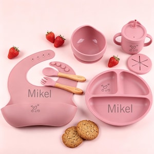 Könnte beinhalten: Ein rosa Baby-Ess-Set mit Lätzchen, Schüssel, geteiltem Teller, Trinkbecher und Besteck. Das Set enthält ein Lätzchen, einen Löffel, eine Gabel und einen Teller mit dem Aufdruck "Mikel". Erdbeeren und Kekse sind ebenfalls enthalten.