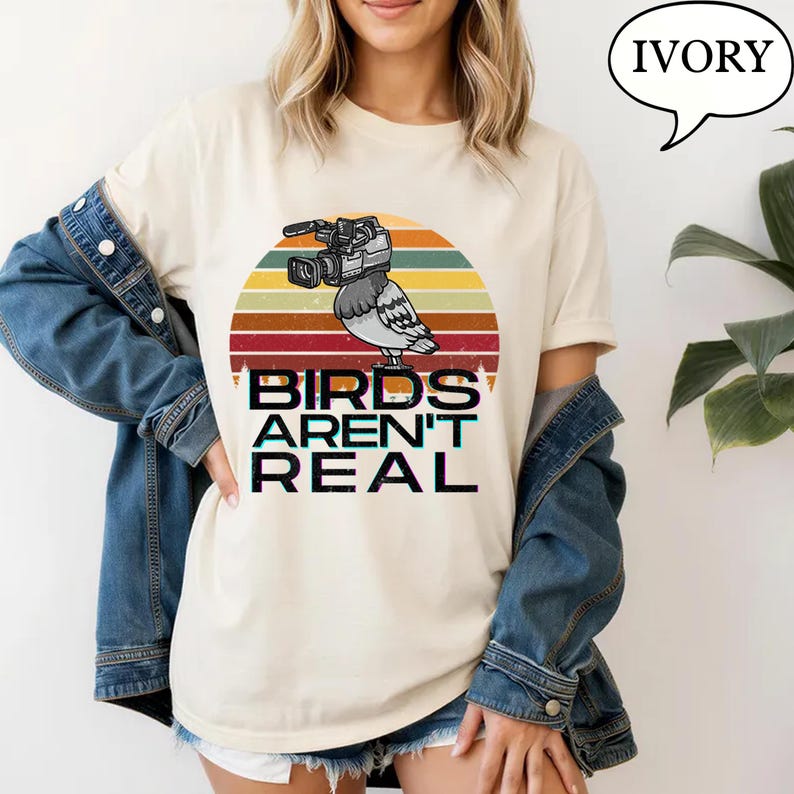 May include: Ivory-colored t-shirt with a graphic of a pigeon wearing a video camera, set against a retro sunset design. The text "BIRDS AREN'T REAL" is printed below the image in bold, black letters. The shirt is a casual wear item.