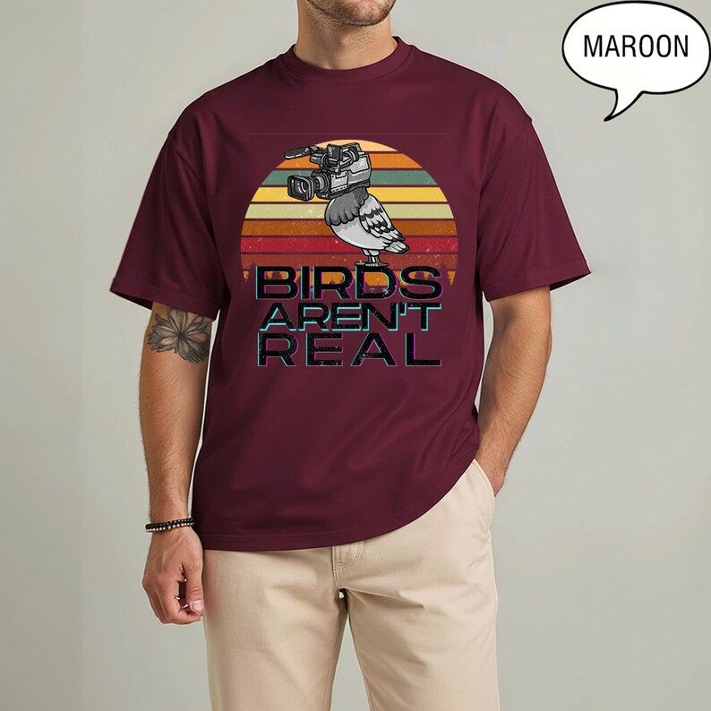 May include: Maroon t-shirt with a graphic of a pigeon wearing a video camera, set against a sunset design. The text "BIRDS AREN'T REAL" is below the pigeon. The shirt is a solid maroon color.