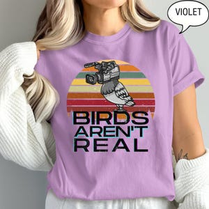 May include: A violet-colored t-shirt features a graphic of a pigeon with a video camera on its head, set against a retro sunset design. The text "BIRDS AREN'T REAL" is printed below the image in a bold, stylized font.