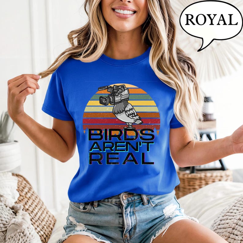 May include: Royal blue t-shirt featuring a graphic of a pigeon with a video camera, set against a retro sunset design. The text "BIRDS AREN'T REAL" is printed below the bird. The shirt is a short-sleeved crew neck.