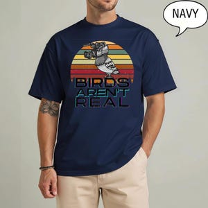 May include: Navy blue t-shirt featuring a retro sunset design with a pigeon wearing a video camera. The text "BIRDS AREN'T REAL" is displayed below the bird. The shirt is a solid navy color.