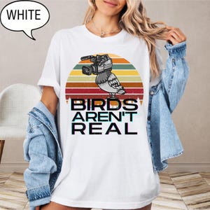 May include: White t-shirt with a graphic of a bird with a video camera, set against a retro sunset design. The text "BIRDS AREN'T REAL" is printed below the bird. A speech bubble with the word "WHITE" is in the upper left corner.