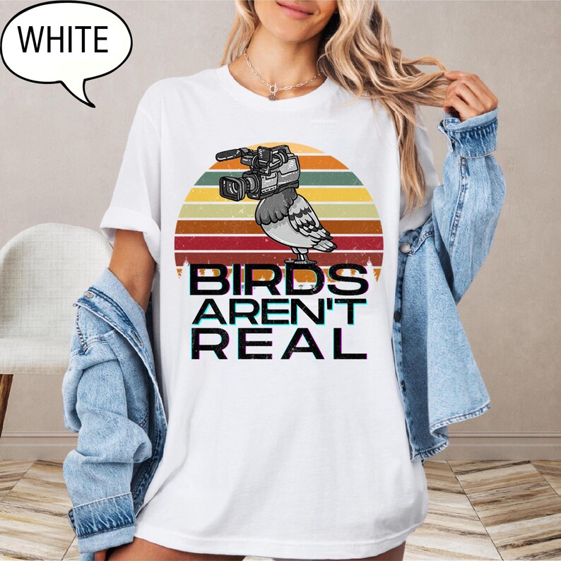 May include: White t-shirt with a graphic of a bird with a video camera, set against a retro sunset design. The text "BIRDS AREN'T REAL" is printed below the bird. A speech bubble with the word "WHITE" is in the upper left corner.