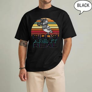 May include: Black t-shirt featuring a graphic of a pigeon with a video camera, set against a sunset design. The text "BIRDS AREN'T REAL" is displayed below the bird. The shirt is a classic crew neck style.