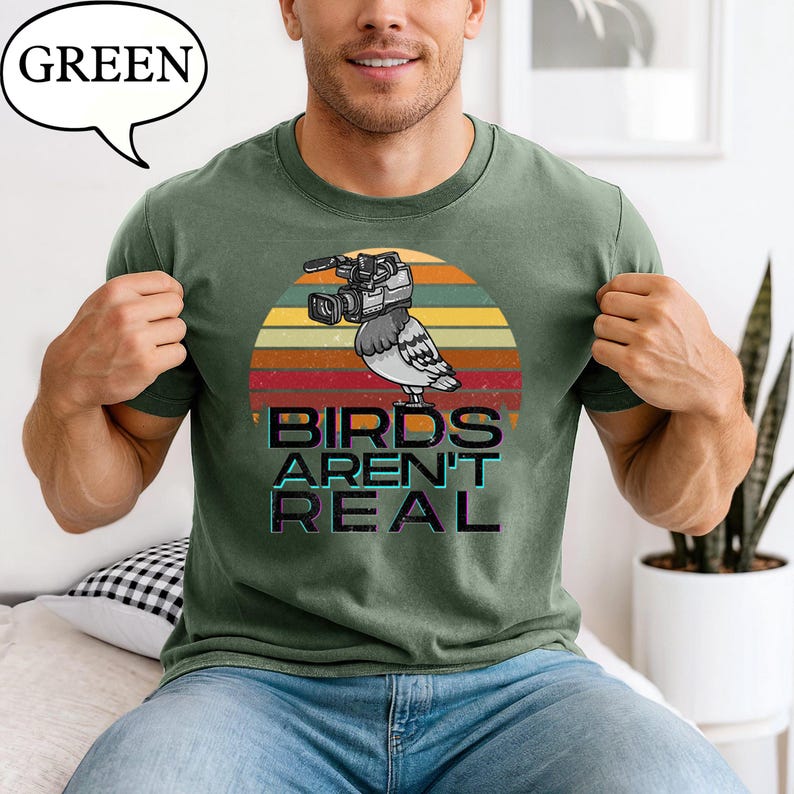May include: Green t-shirt with a graphic of a pigeon wearing a video camera, set against a sunset design. The text "BIRDS AREN'T REAL" is below the image. The word "GREEN" is in a speech bubble in the upper left corner.