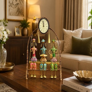 May include: A decorative metal table clock featuring an arched frame with three stylized figures sitting on a bench. The figures are painted in pink, blue, and green, each holding an instrument. A round clock face with black Roman numerals sits at the top.