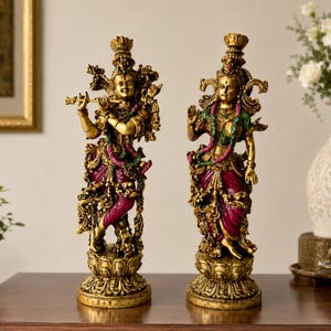 May include: Two ornate gold-toned statues depicting deities standing on lotus bases. The left figure plays a flute, while the right figure has one hand raised. Both feature intricate metallic detailing, magenta and green accents, and elaborate crowns.