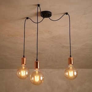 May include: A three-bulb pendant light fitting with Edison-style bulbs. The fixture has a black ceiling mount and black cords. The bulb bases are copper-coloured. The bulbs are round and emit a warm, yellow light.