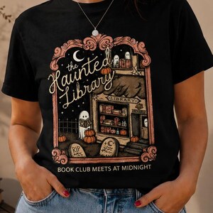 Puede incluir: Una camiseta negra de manga corta con un estampado gráfico de una biblioteca con fantasmas, calabazas y lápidas. El texto dice The Haunted Library y Book Club Meets at Midnight dentro de un marco decorativo con una luna creciente.