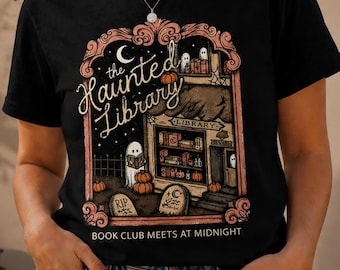May include: A black short-sleeve t-shirt featuring a graphic print of a library building with ghosts, pumpkins, and tombstones. The text reads The Haunted Library and Book Club Meets at Midnight in a decorative frame with a crescent moon above.