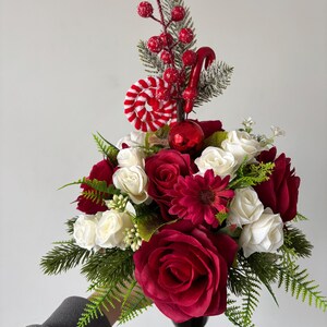 Könnte beinhalten: Ein festliches Blumenarrangement mit dunkelroten und weißen Rosen, einer dunkelroten Margerite und grünen Tannenzweigen. Das Gesteck ist mit roten Beeren, einer rot-weiß gestreiften Zuckerstange, einem roten Wirbelornament und einer roten Kugel.