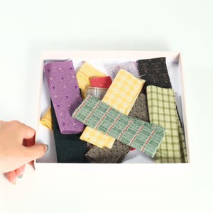 May include: A collection of rectangular fabric swatches in a white box, featuring various patterns and colors including purple with small stars, yellow plaid, green with red stripes, black with dots, and olive green checkered designs.
