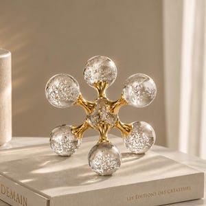 May include: A decorative starburst sculpture with six clear glass orbs filled with glitter, connected by a gold-colored metal framework. The sculpture sits atop a beige book with the text "A DEMAIN".