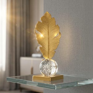 May include: A gold-colored decorative feather sculpture. The feather sits atop a clear glass orb with bubbles, mounted on a gold square base. The sculpture is displayed on a glass shelf.