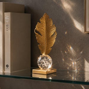 May include: A gold-colored feather-shaped decorative object with a clear glass orb base, resting on a square gold base. The object is displayed on a glass shelf with books and a marble block in the background. The books have titles like "Live Beautifully".