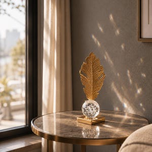May include: A gold-colored feather sculpture atop a clear glass orb with decorative elements, mounted on a square gold base. The sculpture sits on a round table with a marble-like top and gold trim.