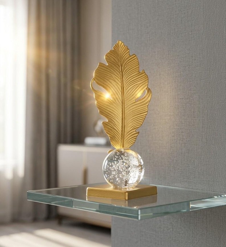 May include: A gold-colored decorative feather sculpture. The feather sits atop a clear glass orb with bubbles, mounted on a gold square base. The sculpture is displayed on a glass shelf.