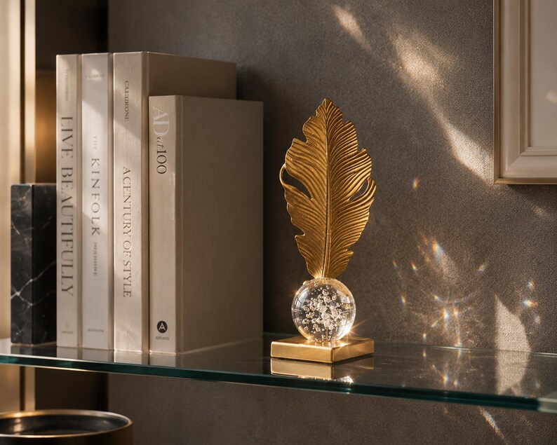 May include: A gold-colored feather-shaped decorative object with a clear glass orb base, resting on a square gold base. The object is displayed on a glass shelf with books and a marble block in the background. The books have titles like "Live Beautifully".