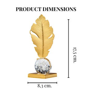 May include: A gold-colored decorative feather sculpture. The feather is mounted on a clear globe filled with silver glitter, which sits atop a gold square base. The sculpture is 17.5 cm tall and 8.3 cm wide.