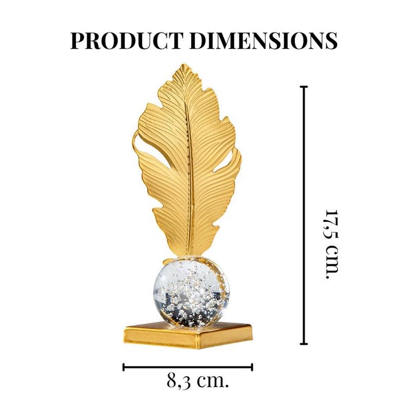 May include: A gold-colored decorative feather sculpture. The feather is mounted on a clear globe filled with silver glitter, which sits atop a gold square base. The sculpture is 17.5 cm tall and 8.3 cm wide.