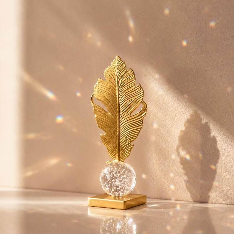 May include: A gold-colored decorative feather sculpture. The feather is detailed with intricate lines and sits atop a clear glass orb filled with small bubbles. The orb is mounted on a square gold base. The sculpture is displayed against a neutral background.