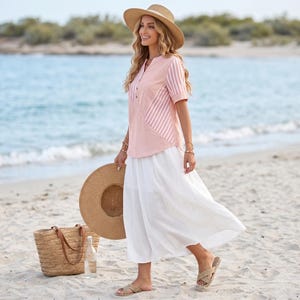 May include: A short-sleeved blouse featuring a solid light pink center panel and vertical white and pink striped side panels, paired with a long, flowing white skirt. The outfit includes a wide-brimmed straw hat and woven straw tote bag.