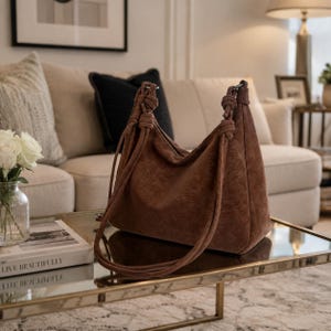 Può includere: Una borsa a spalla in camoscio marrone con cinturini annodati è appoggiata su un tavolino con piano in vetro. La borsa ha un design morbido e un aspetto morbido e strutturato. Lo sfondo include un divano color crema e oggetti decorativi.