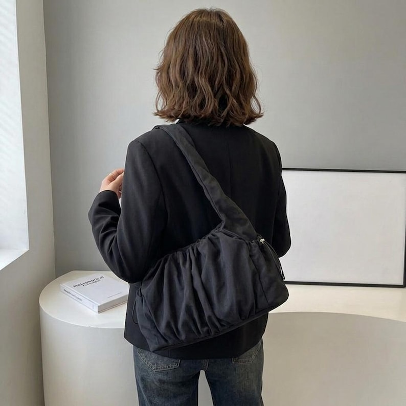 以下が含まれることがあります： ギャザーデザインの黒いショルダーバッグ。バッグは肩からかけられ、長いストラップが付いています。人物は黒いブレザーとブルージーンズを着用しています。バッグは柔らかい、おそらくナイロン素材で作られているようです。