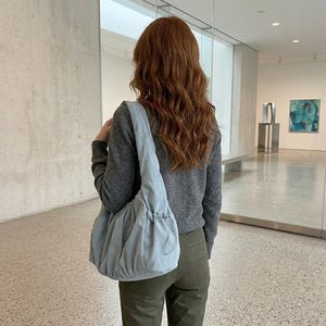 以下が含まれることがあります： ギャザーデザインのライトブルーのショルダーバッグ。バッグは柔らかい素材でできており、長いストラップが付いています。バッグは肩にかけています。その人はグレーのセーターとグリーンのパンツを着ています。
