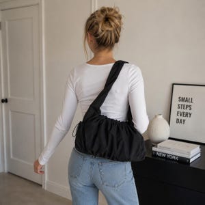 以下が含まれることがあります： 肩にかけられた、ギャザーデザインの黒いショルダーバッグ。 バッグは柔らかい素材でできており、巾着開閉式です。 画像には、白い長袖シャツとブルージーンズも写っています。 額入りのプリントには「SMALL STEPS EVERY DAY」と書かれています。