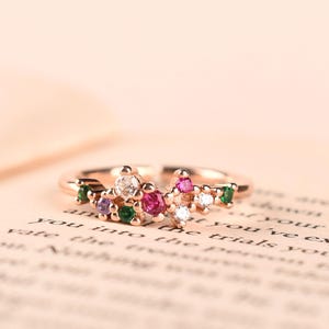 Peut inclure: Une bague de couleur or rose avec un design à anneau ouvert orné d'un groupe de petites pierres précieuses multicolores, incluant des pierres transparentes, roses, violettes et vertes disposées en un motif décoratif et asymétrique.