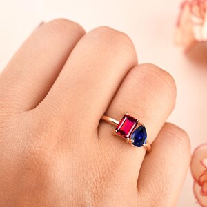 May include: A gold ring featuring two prominent gemstones: a rectangular emerald-cut red stone and a pear-shaped blue stone. The stones are set side-by-side on a thin, polished gold band, creating a distinct contrast between the red and blue colours.