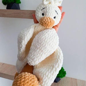 Pode incluir: Um brinquedo de pel&uacute;cia em croch&ecirc; com formato de ganso, feito de fio branco macio com acabamento texturizado e irregular. O ganso apresenta uma cabe&ccedil;a arredondada com olhos bordados em preto, um bico bege e p&eacute;s bege, com um pequeno tufo de fio.