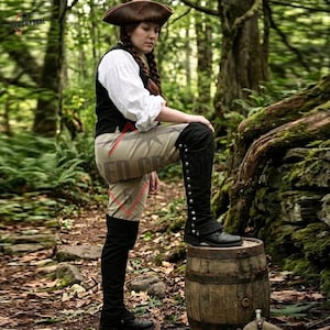 May include: A brown tricorn hat, a white long-sleeved shirt, a black vest, beige trousers, and black over-the-knee boots with silver-toned buttons along the side. The outfit is worn by a person with long braided hair standing in a forest.