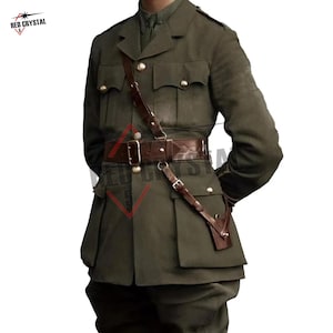 May include: An olive drab military-style tunic featuring a structured collar, long sleeves, and four front pockets with button closures. A brown leather Sam Browne belt with a diagonal shoulder strap and metal buckles is worn over the jacket.