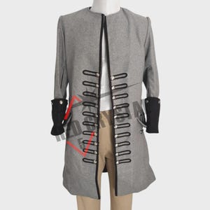 Peut inclure: Un manteau gris à manches longues avec une encolure haute sans col et une ouverture frontale ornée de deux rangées verticales de boucles tressées noires et de boutons en métal argenté. Les manches ont des poignets noirs contrastants sur un pantalon beige.