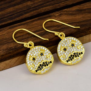 May include: A pair of gold-tone drop earrings featuring circular pendants. Each pendant is encrusted with clear white stones and accented with black stones arranged to form a mustache and eye pattern, suspended from simple gold-tone ear wires.