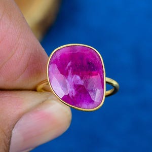May include: A gold ring featuring a large, irregular, faceted magenta gemstone. The stone has a polished surface with visible internal inclusions and a vibrant pink hue, set within a simple gold band that encircles the finger.