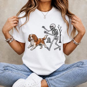 Op de afbeelding: Een wit t-shirt met een grafische print van een bruin-witte Cavalier King Charles-spaniël die voor een skelet uit rent. Het skelet is afgebeeld met uitgestrekte armen bij een grafsteen en het silhouet van een kale boom op het shirt.