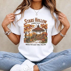 Könnte beinhalten: Ein weißes Kurzarm-T-Shirt mit einer Grafik eines Pavillons, umgeben von herbstlichen Bäumen und Kürbissen. Der Text lautet Stars Hollow Connecticut und Where You Lead I Will Follow in brauner Schrift. Das Design enthält herbstliche Farben.