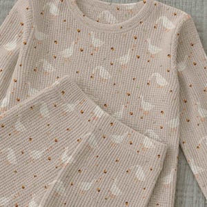 Puede incluir: Un conjunto de camisa de manga larga y pantalones en tejido de punto gofrado beige con un estampado de pequeñas ocas blancas y diminutas bellotas marrones. La tela presenta un patrón de rejilla texturizado en toda la prenda.