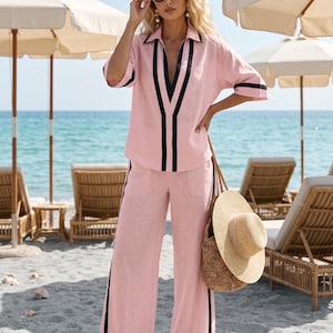 Op de afbeelding: Een lichtroze linnen tweedelige outfit met zwarte bies. De set bevat een overhemd met kraag en een broek met wijde pijpen. Een strooien totebag en een zonnehoed zijn ook zichtbaar. De outfit wordt gedragen op een zandstrand met ligstoelen en parasols op de achtergrond.