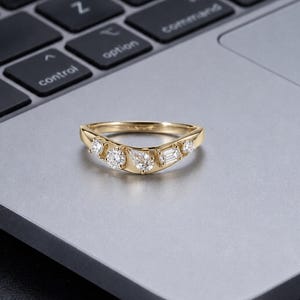 May include: A gold chevron-shaped ring featuring five clear, faceted stones set along the band. The stones include two small round cuts on the ends, a larger round cut, a pear-shaped stone, and a rectangular baguette-cut stone in the center.