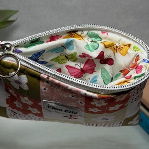 May include: A zipped pouch featuring a patchwork exterior with floral patterns in shades of olive green, pink, and white. A rectangular label with the text "Ehm Elle" is stitched on the front. The interior lining displays a colourful butterfly print.