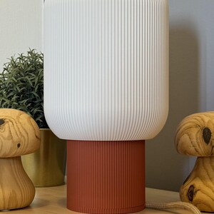 Pode incluir: Uma moderna luminária de mesa com uma cúpula branca e canelada e uma base cor de ferrugem. A luminária é cilíndrica e está sobre uma mesa de madeira. Estatuetas decorativas de cogumelos de madeira e uma pequena planta também estão presentes.